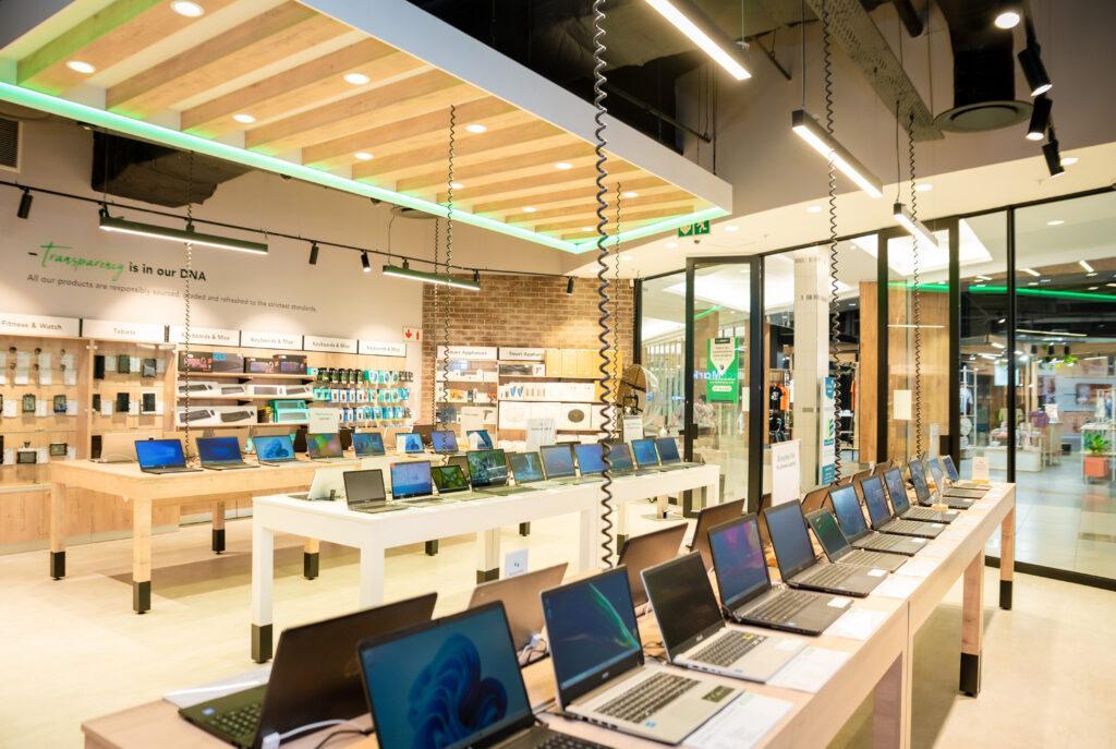 Modern electronics store interior showcasing pre-owned and new tech products, including laptops displayed on tables, shelves stocked with cellphones, tablets, and accessories, under sleek lighting and a banner emphasizing sustainability and transparency.